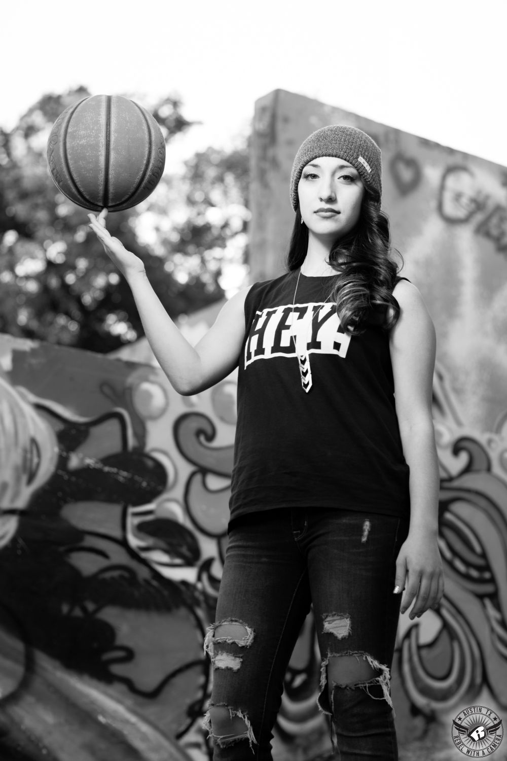 Dramatic black and white image of urban looking high school senior girl with hair and makeup by Divaz Fabula  spinning a basketball at Castle Hill Graffiti Wall in Austin, Texas, taken by Austin senior portrait photorapher Rebel with a Camera.