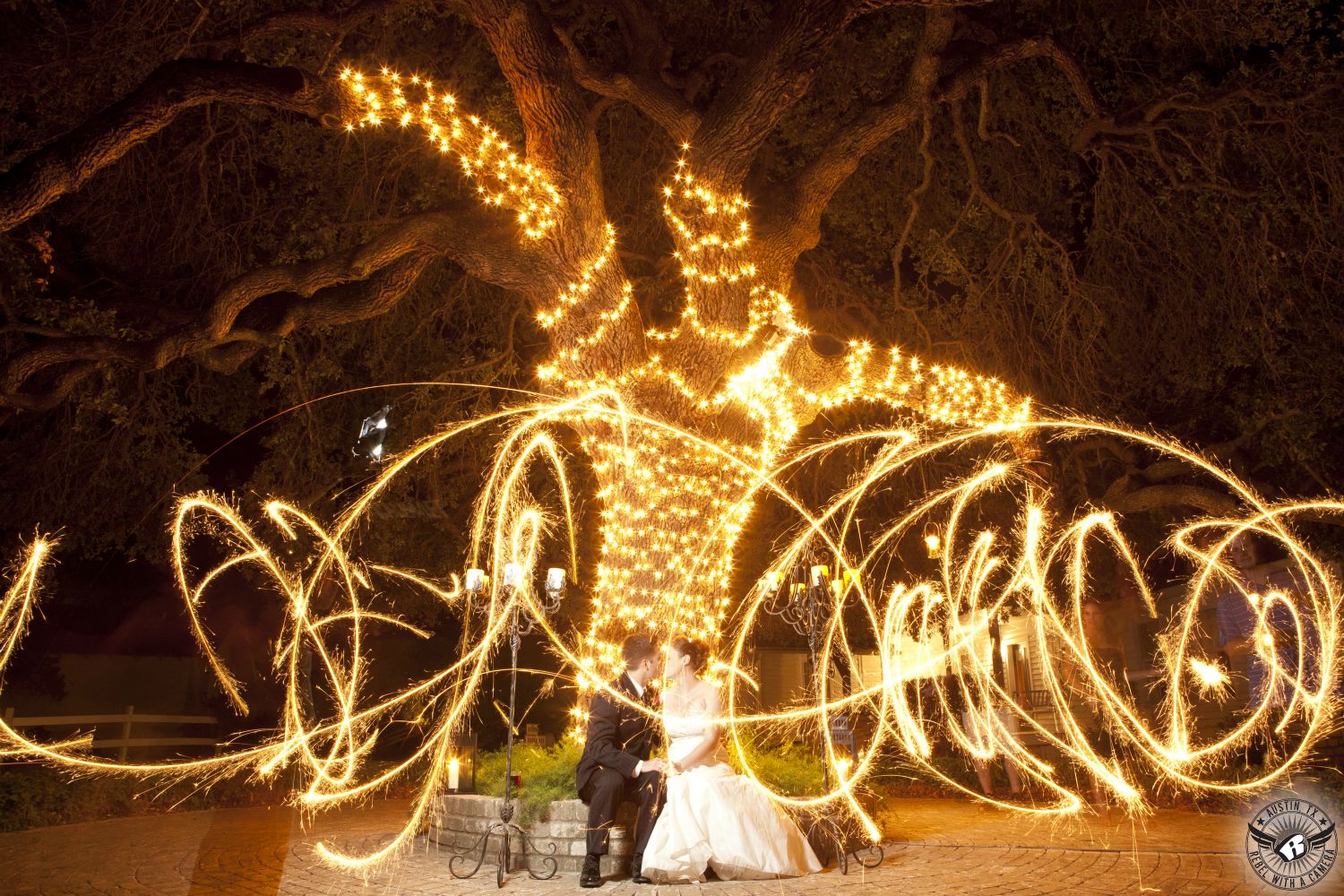 Bride and groom kiss at night during their wedding in front of oak tree with christimas lights and sparklers swirling around at Inn at Salado.
