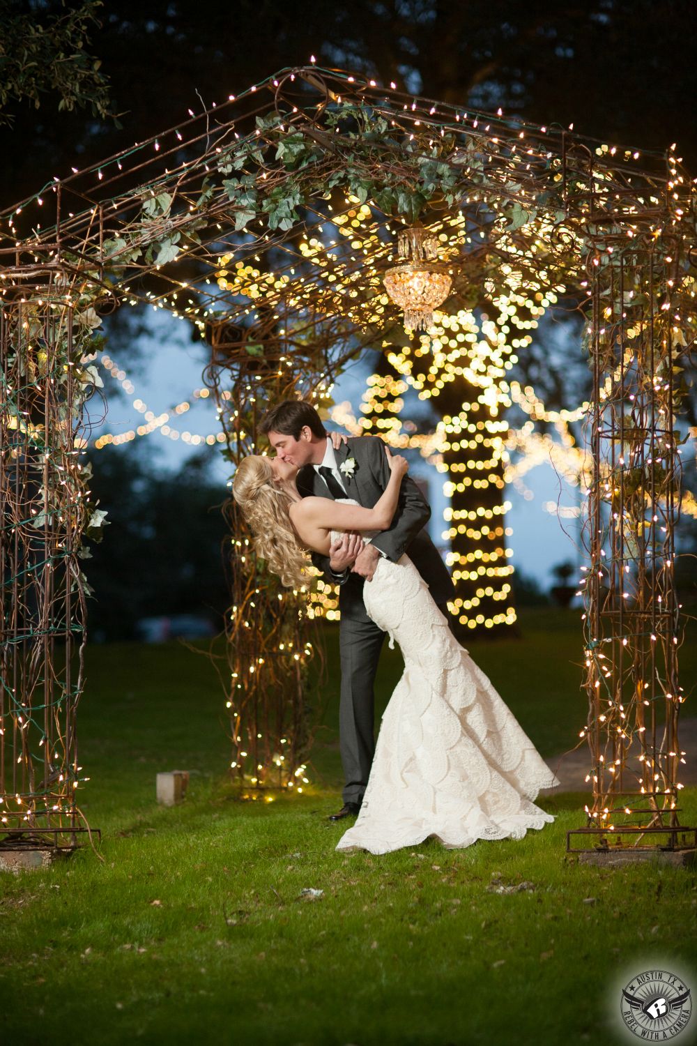 Groom dips bride and kisses her in the twilight in front of the arbor at Kindred Oaks covered by twinlky lights just after the wedding.