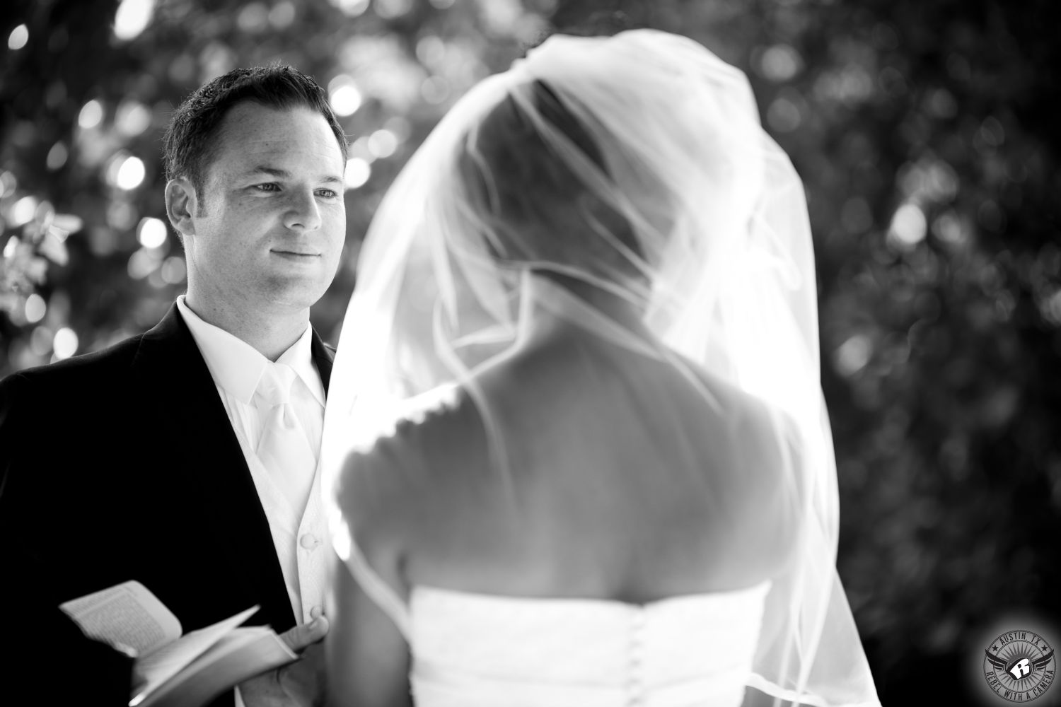 Black and white image of groom looking at bride with love as he says his vows during outdoor wedding ceremony at the arbor at Nature's Point wedding venue on the shores of Lake Travis.