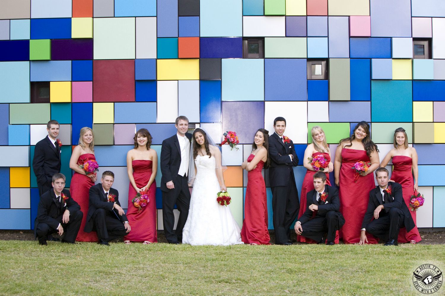 Bridal party with bridesmaids in strapless red full length dresses and orange and pink bouquets stand in front of bright and colorful mosiac wall at Discovery Green Park in Houston, Texas, during the wedding.
