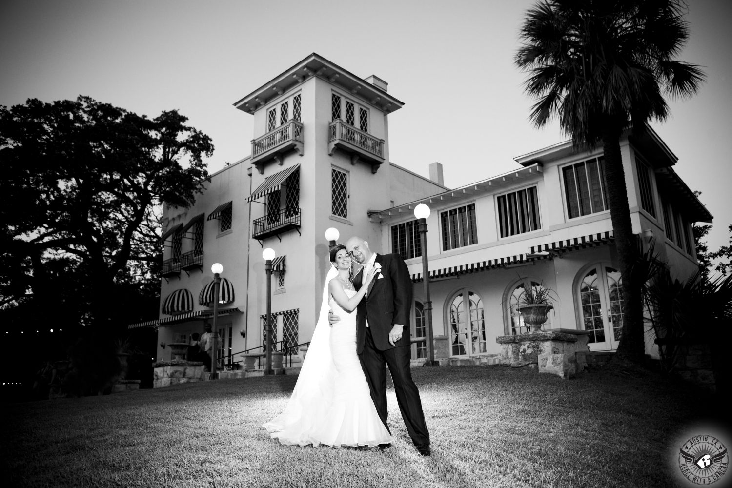 Picture of bride in glamorous strapless bridal gown and floor length veil with groom in black tuxedo and white tie standing in front of the mansion at Laguna Gloria Austin wedding venue.