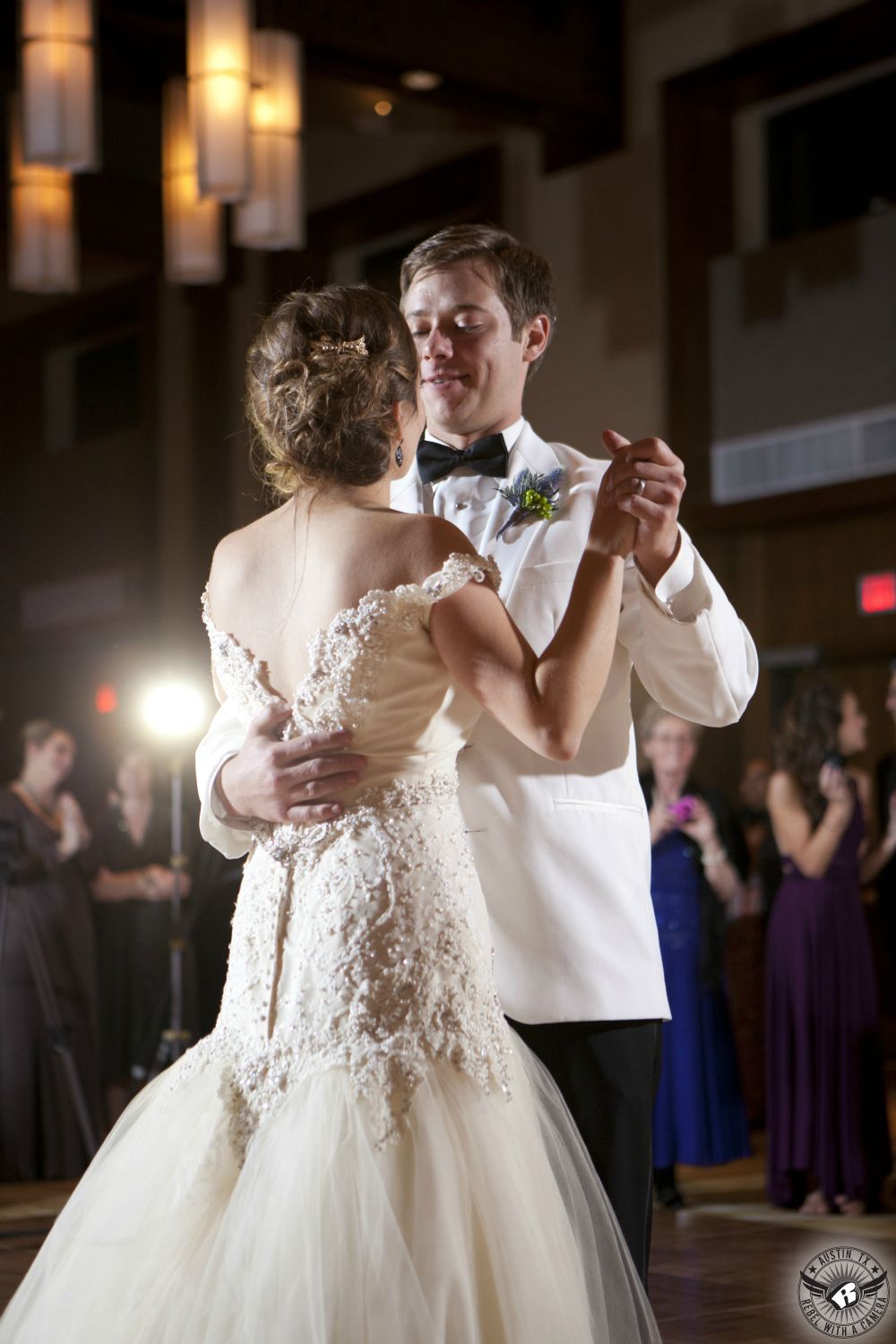 Bride in sparkly off the shoulder bridal gown and groom in white tuxedo jacket and black bowtie with David Kurio floral boutonniere first dance at the AT&T Executive Education and Conference Center ballroom with live jazz band at their wedding reception.