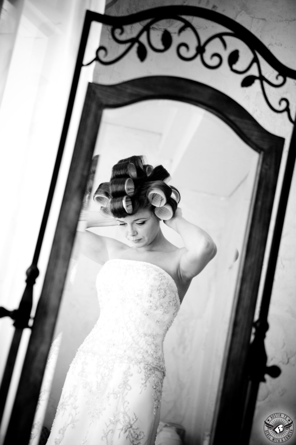 Black and white image of beautiful bride in curlers and elegant strapless bridal gown embellished with gold embroidery adjusts her hair in the mirror at Villa Antonia Tuscan styled Austin wedding venue.