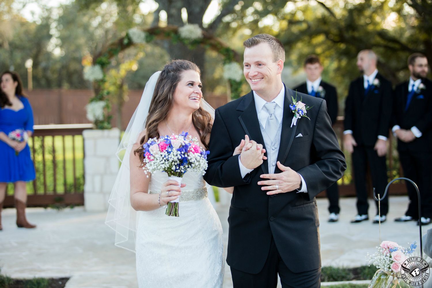 Happy brunette bride with pink and royal blue bouquet walks down the aisle after the wedding ceremony with her room in black tuxedo adn grey vest and tie as new husband and wife at Gabriel Springs Event CenterAaustin area wedding venue.