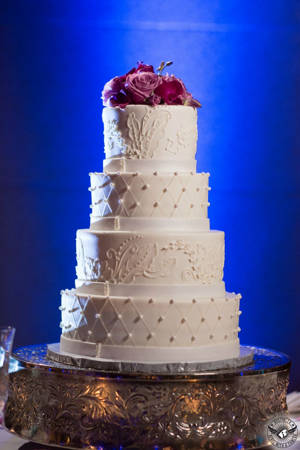 Four tier white bridal wedding cake by The Cake Plate in Austin at the Grand Ballroom at 1900 University Avenue coordinated by Barbara's Brides with royal blue uplighting and magenta floral cake topper by Wendee Sawran Austin event designer at wedding reception.
