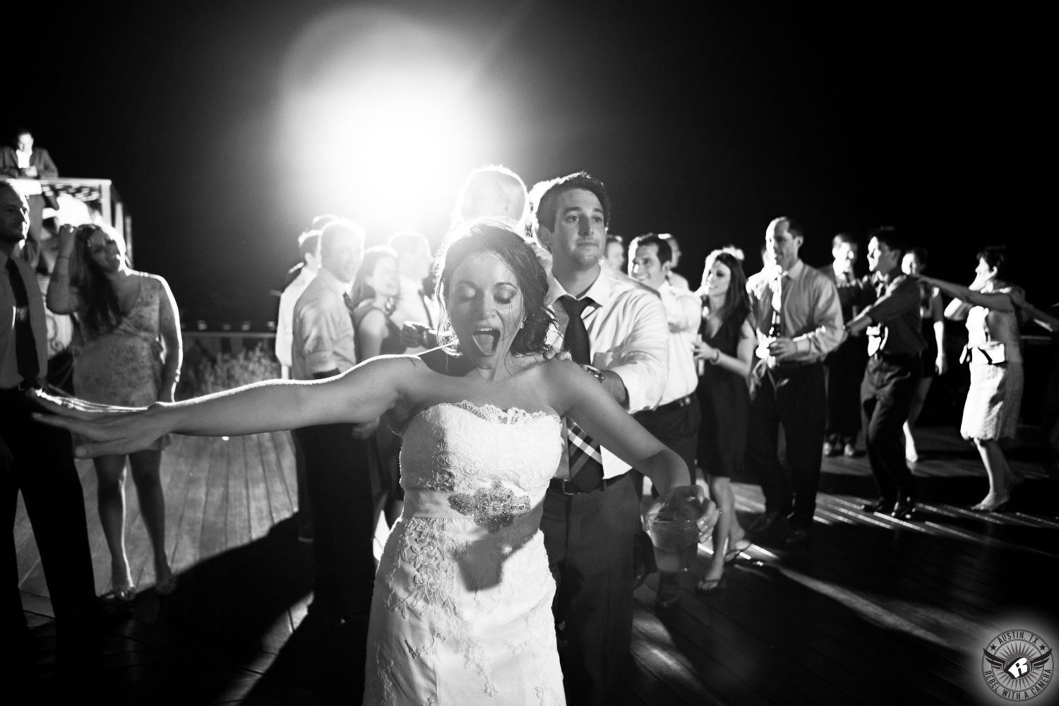 Black and white image of wild dancing conga line on the deck over Lake Travis with the bride in front during wedding reception at Nature's Point Austin wedding venue.