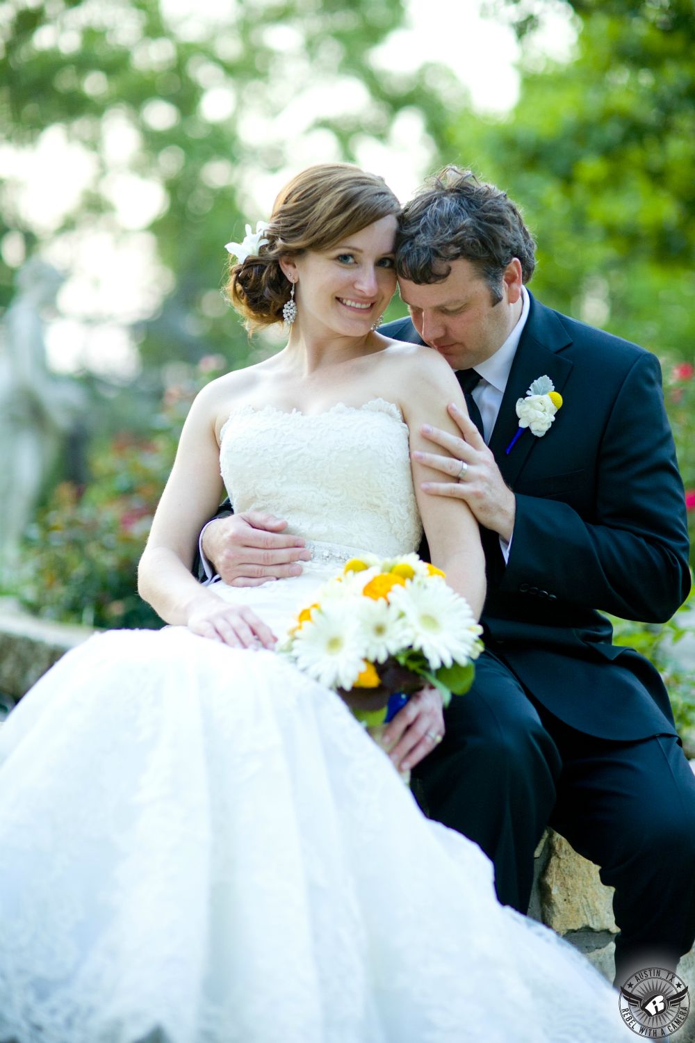Groom kisses shoulder of bride in white lace strapless bridal down with sparkly sash who smiles at the camera and holds a yellow billy balls and daisy bridal bouquet by Bouquets of Austin at Nature's Point Austin wedding venue on the shores of Lake Travis.