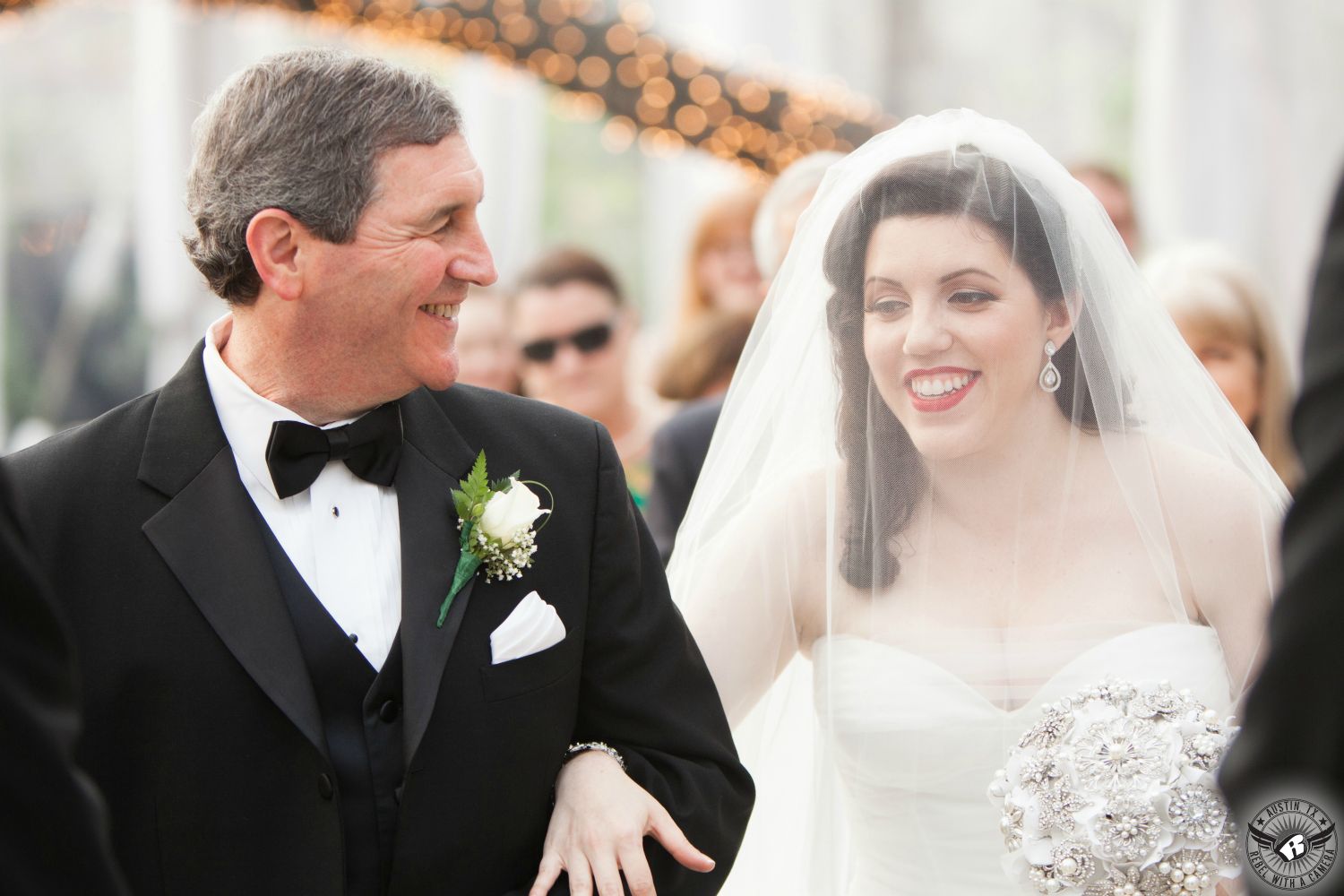 Beautiful brunette bride in blusher veil is walked down the aisle by her loving father in a black tuxedo with a black bowtie at wedding ceremony in the winter tent at the Allan house wedding venue in downtown Austin.