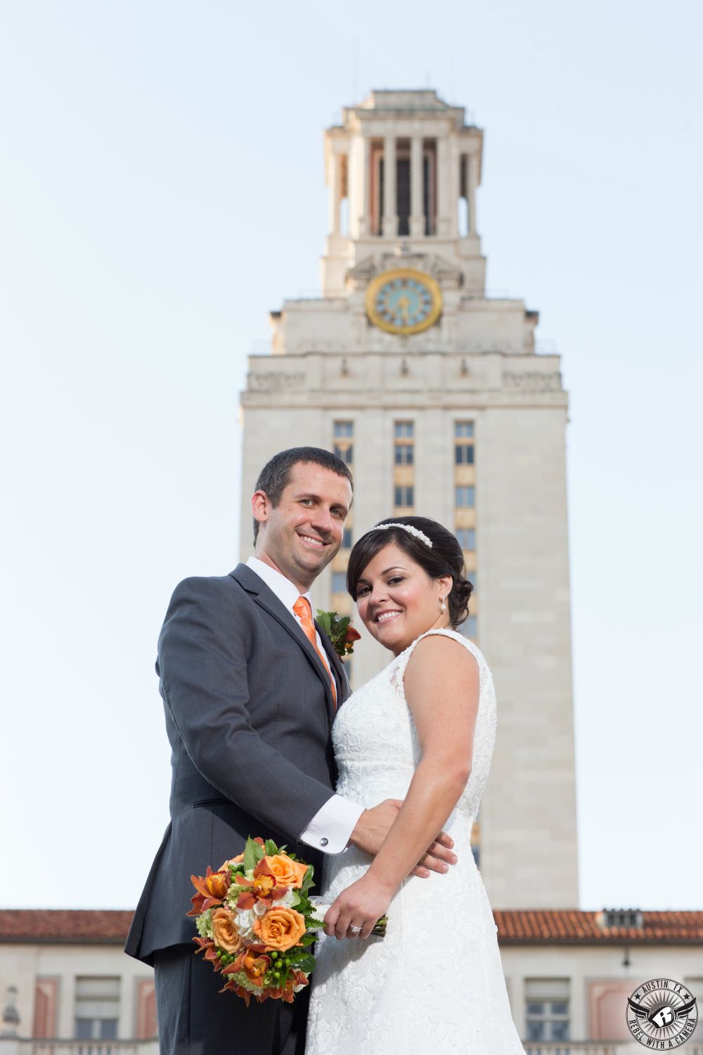 Bride with orange orchid bouquet by Fancy Florals Austin wedding florist and groom in grey suit on their wedding day in front of the UT tower at the University of Texas campus.