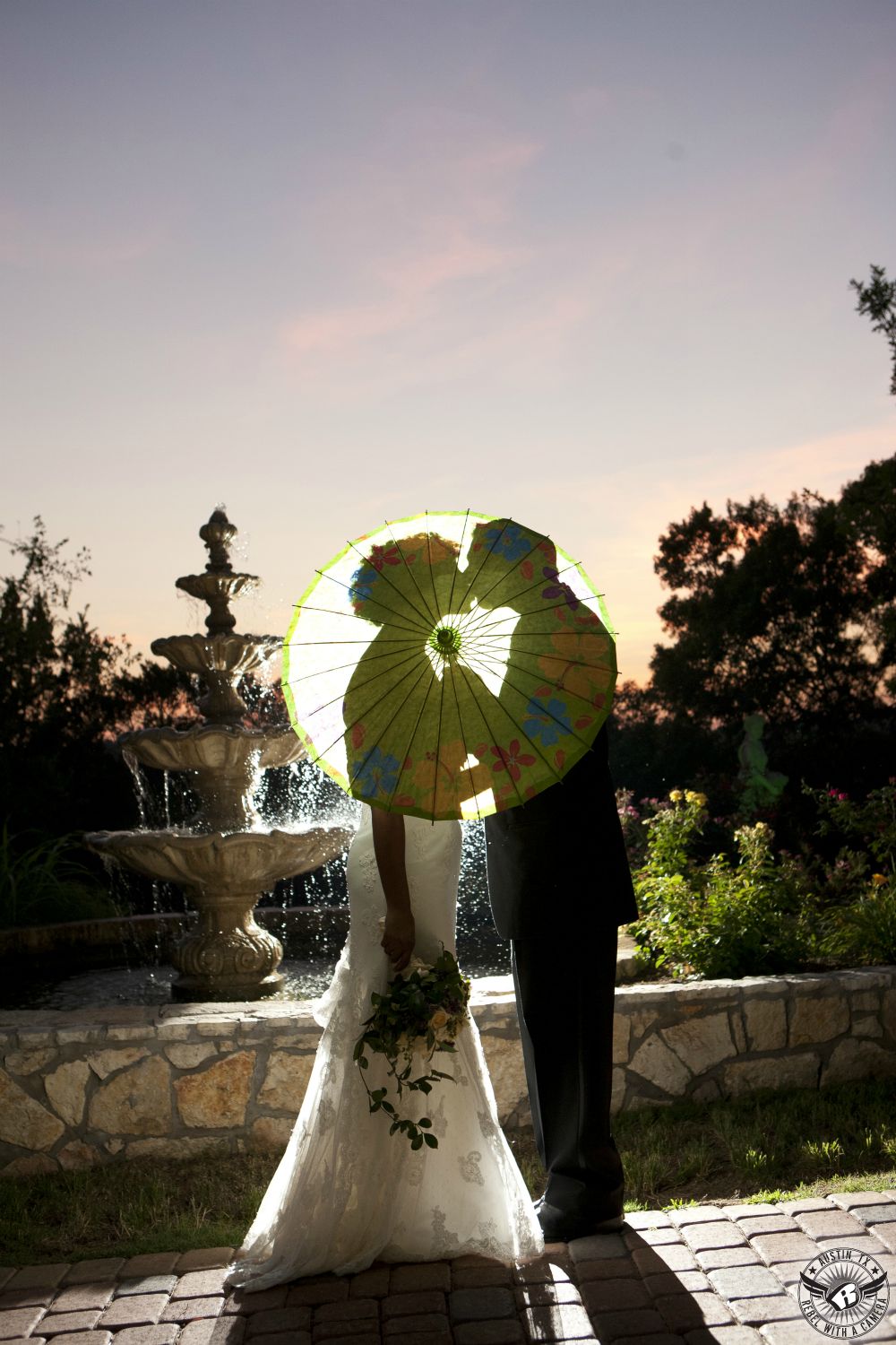 Silhouette of bride with exquisite bouquet by Visual Lyrics Floral Artistry Austin wedding florist and groom with brightly colored parasol  in front of fountain with sunset on their Austin wedding at Nature's Point on the shores of Lake Travis.