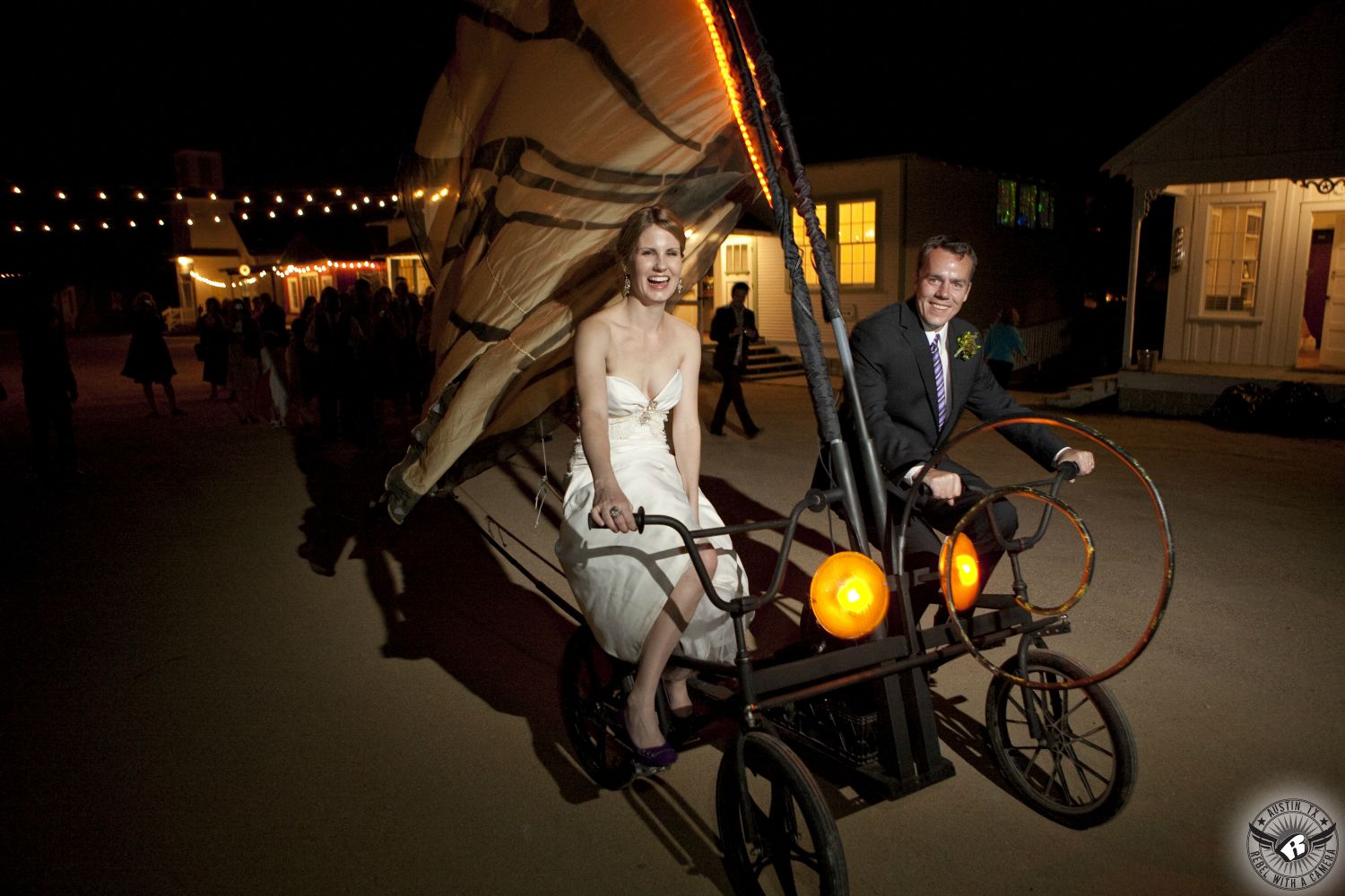 Blonde bride in elegant  strapless wedding dress and groom exit the wedding reception on a wild and whimsical orange butterfly bicycle built for two at Star Hill Ranch Austin area wedding venue.