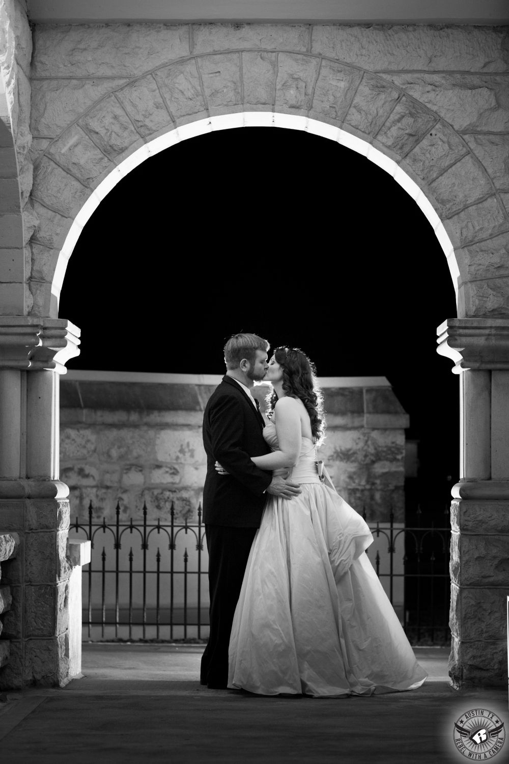 Dramatic black and white image of bride and groom standing under limestone arch with wrought iron fence in the background at night at Chateau Bellevue wedding venue in Austin.