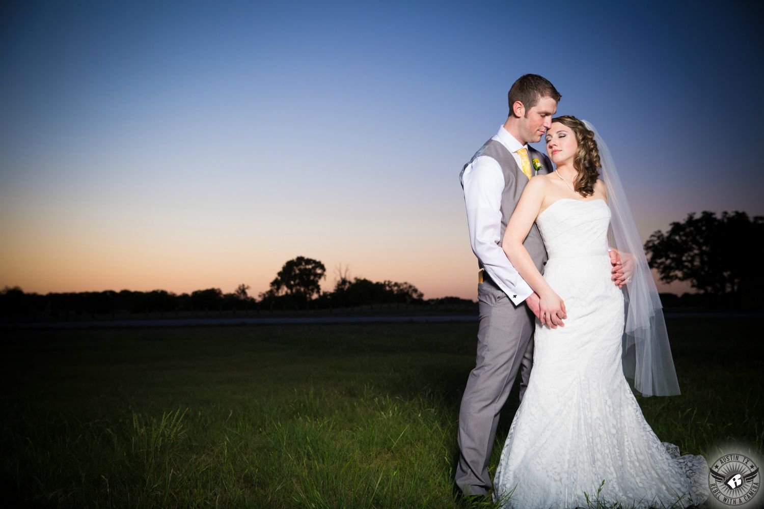 Beautiful brunette bride in strapless lace wedding dress and groom in grey vest at Texas Old Town wedding venue embracing in front of a gorgeous sunset standing in a field.