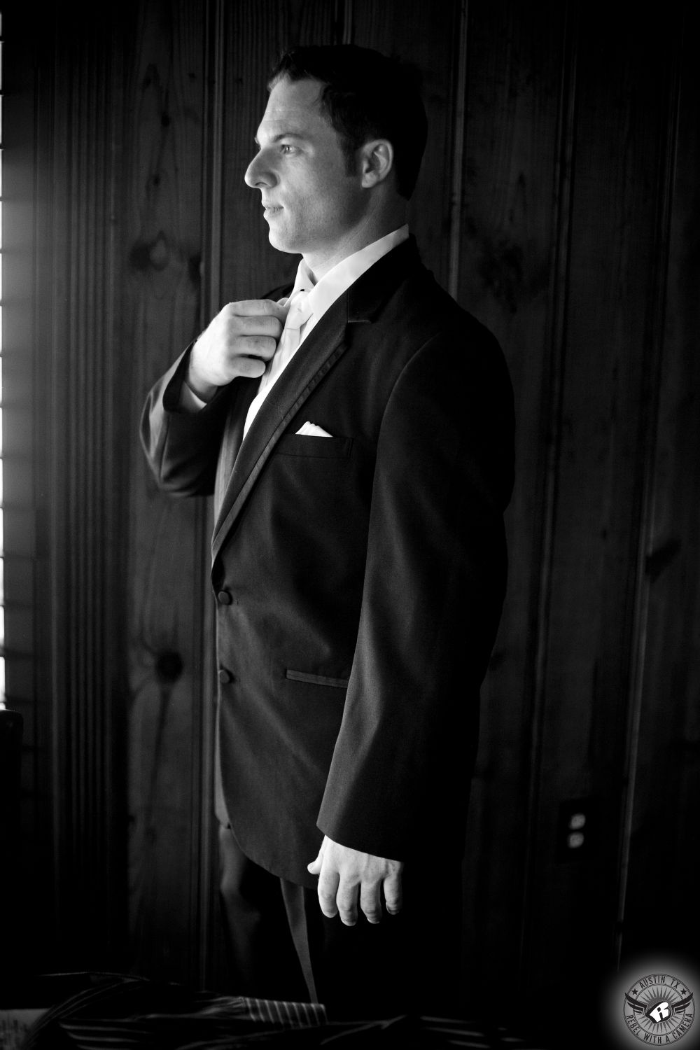 Dramatic black and white image of handsome groom straightening his tie in the window light in the groom's cottage on his wedding day at Nature's Point Austin wedding venue on the shores of Lake Travis.