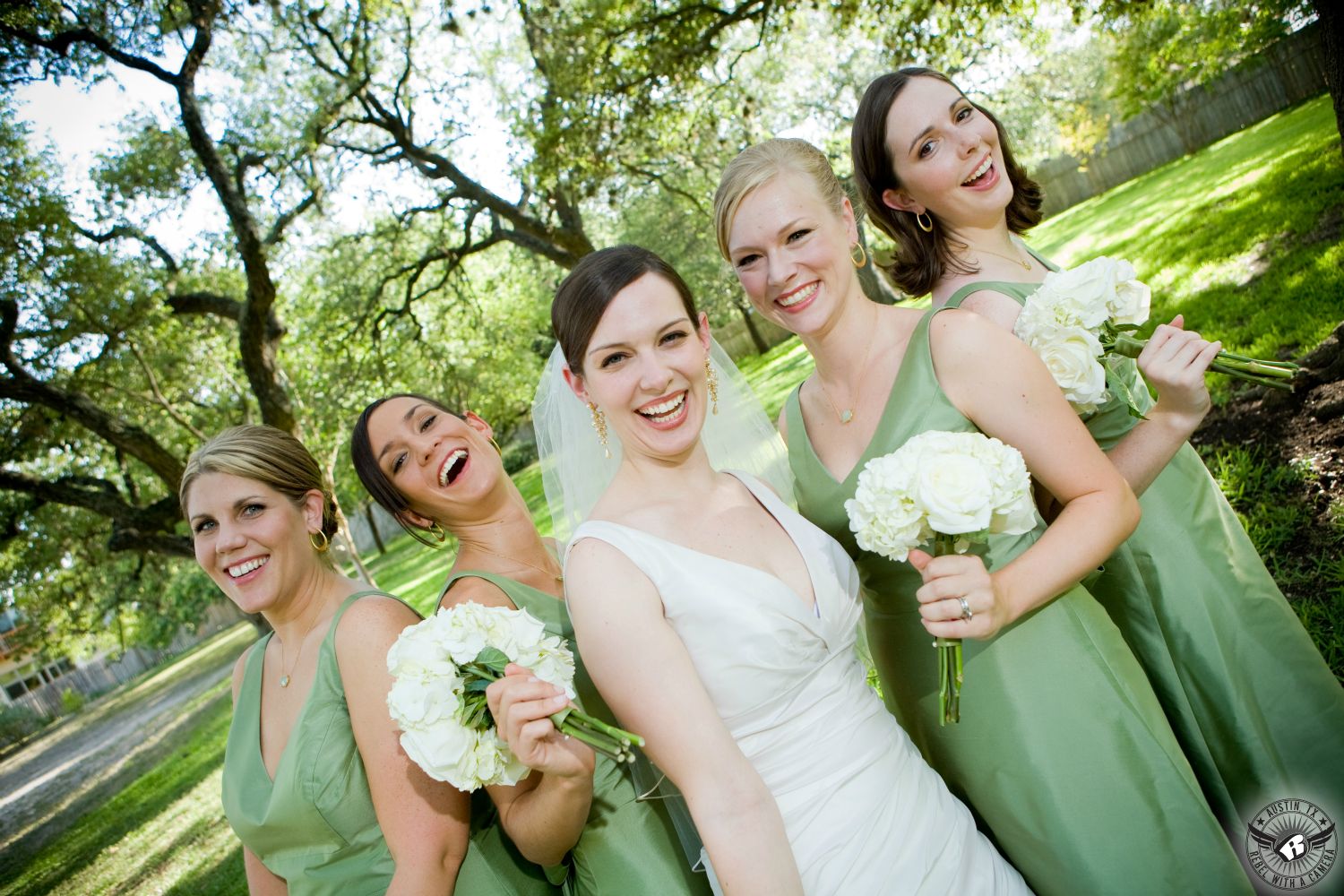 Happy bride and bridesmaids in sea green bridesmaid dresses and with white hydrangea bouquets under oak trees at Green Pastures Austin wedding venue.