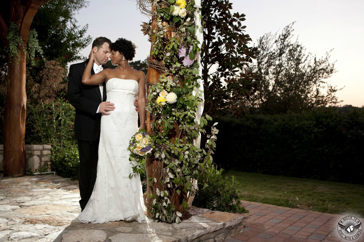 Bride in strapless white wedding dress with exquisite bouquet by Visual Lyrics Floral Artistry Austin wedding florist and groom in black tuxedo embrace under the wedding ceremony arbor at Nature's Point Austin wedding venue on the shores of Lake Travis.
