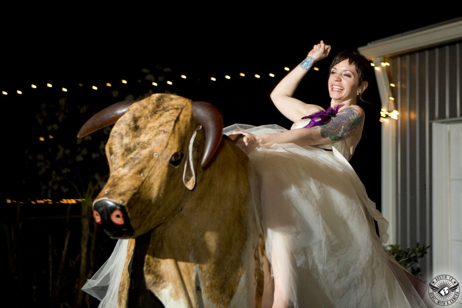 Wedding picture of a wild bride wearing a custom designed haute couture wedding dress riding on a mechanical bull at The Phoenix wedding venue in Columbus, Texas.