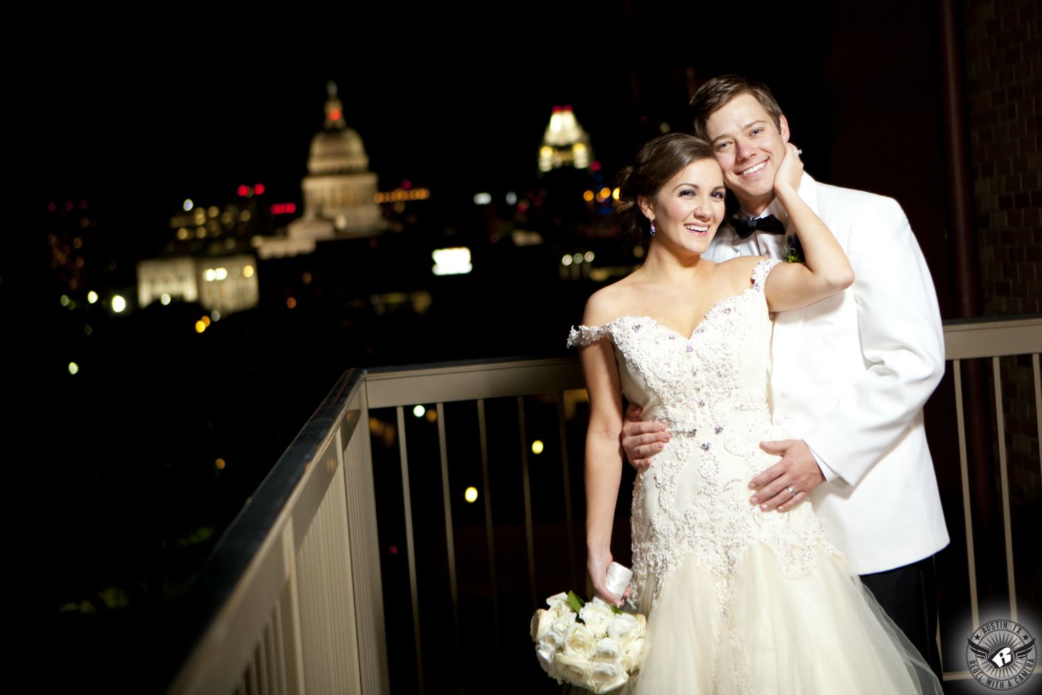 Wedding picture of the bride with white David Kurio bridal bouquet and groom on the balcony of theAT&T Executive Education and Conference Center on the UT on the UT campus at night with a view of the Texas State Capitol building taken by Austin wedding photographer.