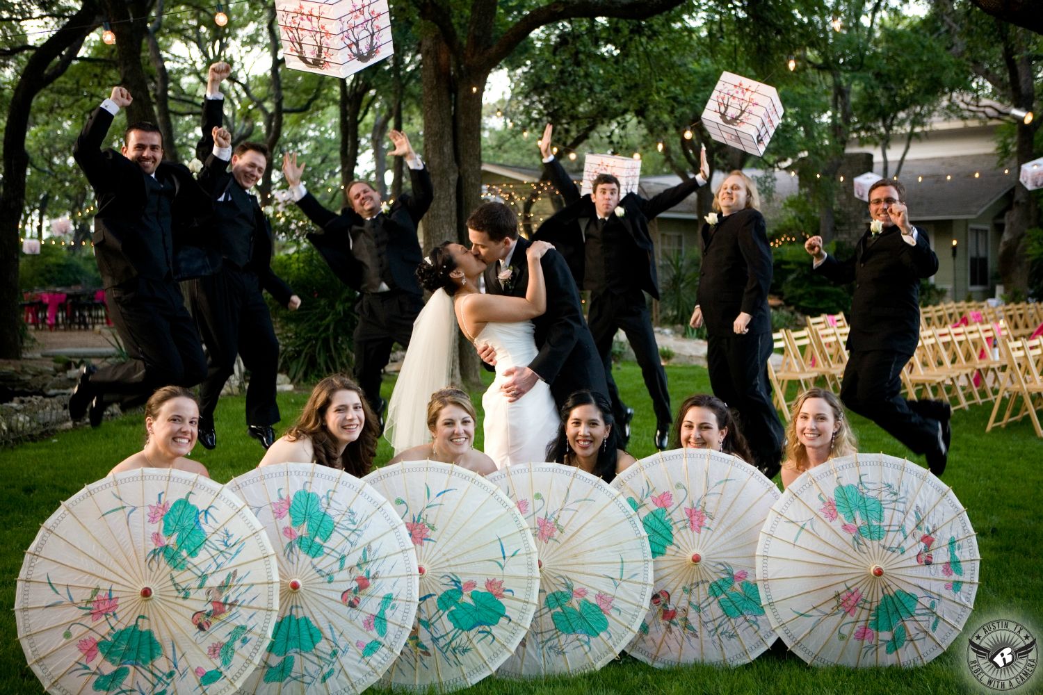 Fun wedding party picture where bride and groom are kissing groomsmen are jumping and bridesmaids are holding colorful parasols and colorful square paper lanterns at Vista at Seward Hill Austin wedding venue taken by Austin wedding photographer.