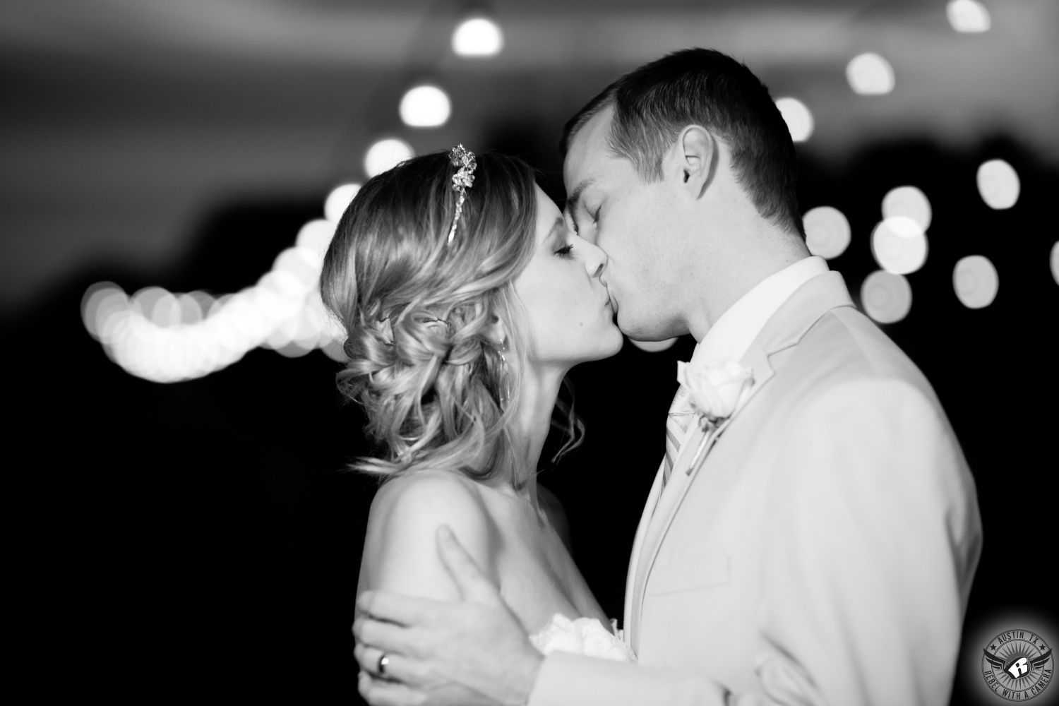 Black and white image of bride with updo and groom kiss in front of twinkly lights with lots of bokeh at Nature's Point Austin wedding venue taken by Austin wedding photographer.