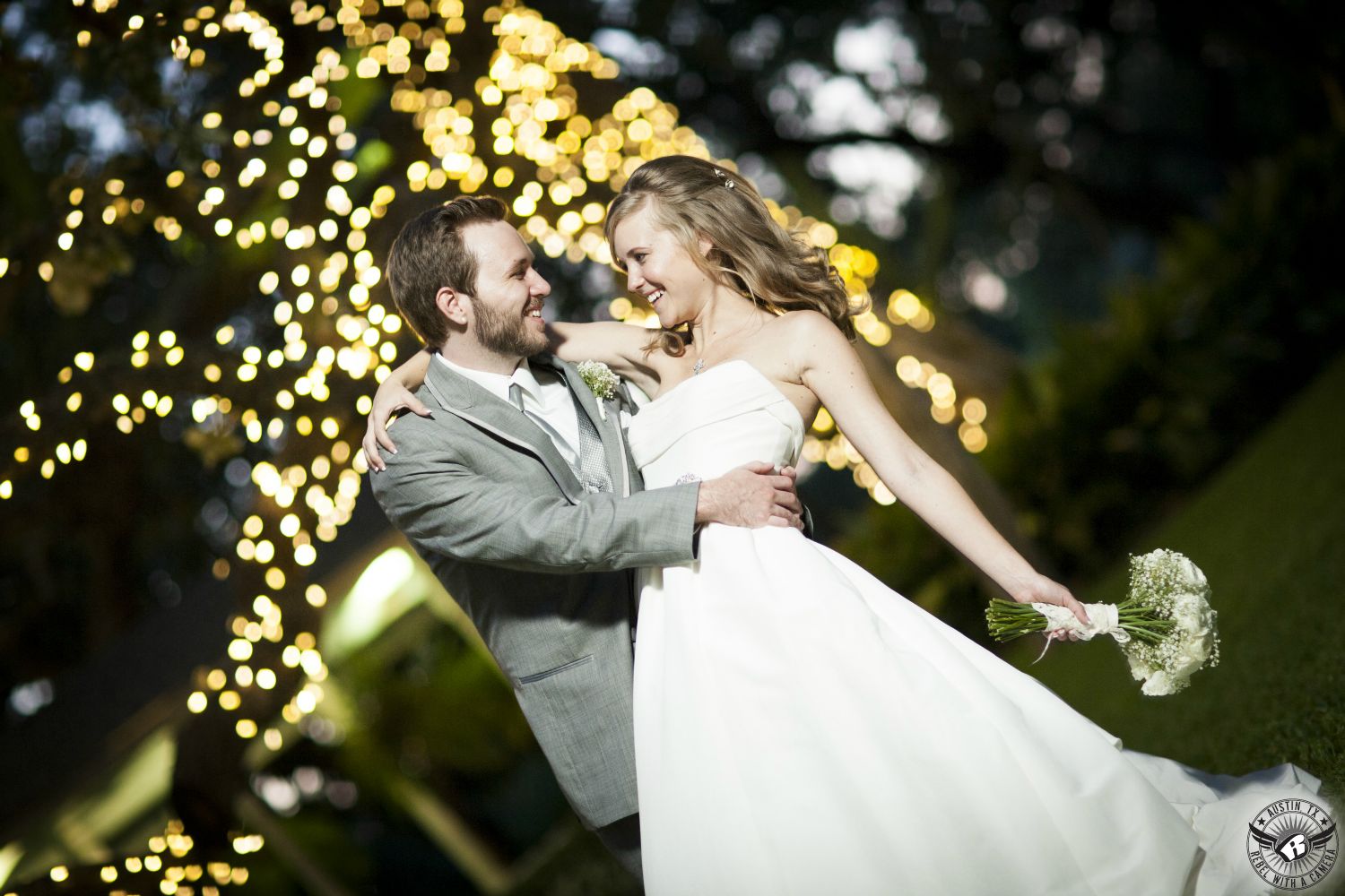 Picture of groom in grey suit dipping bride in strapless dress in front of twinkly lights on tree at Green Pastures Austin wedding venue by Austin Wedding photographer.