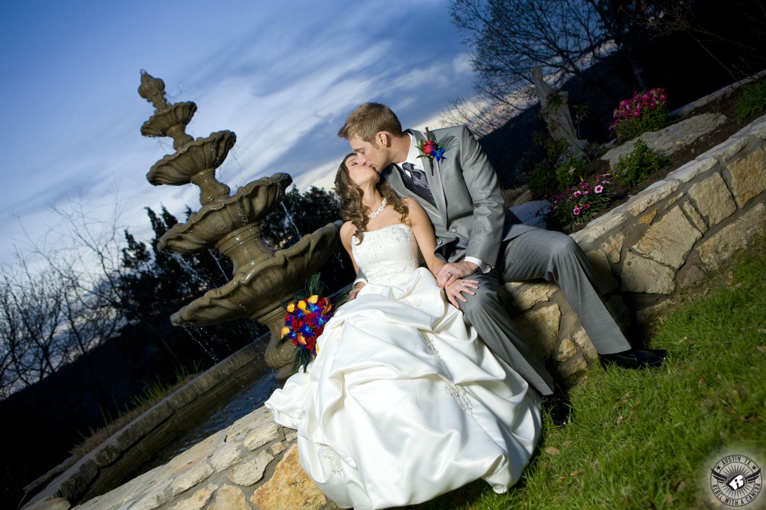 Bride in gorgeous ruffled wedding ballgown and groom in grey suit kissing in front of fountain with beautiful sky at Austin wedding venue Nature's Point on the shores of Lake Travis taken by Austin wedding photographer.