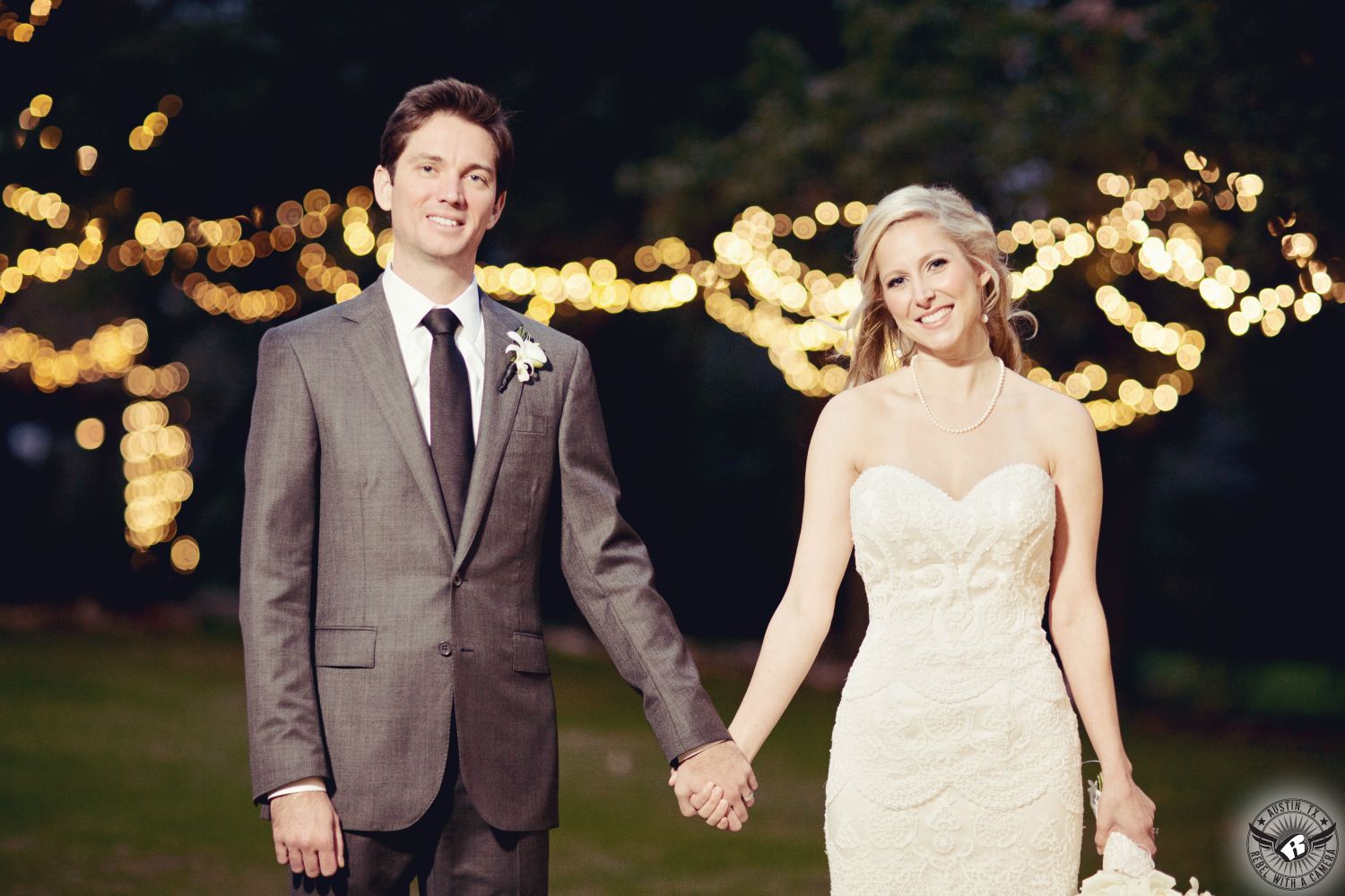 Happy wedding couple holds hands in front of twinkly lights in the trees at Kindred Oaks Austin wedding venue picture taken by Austin wedding photographer.