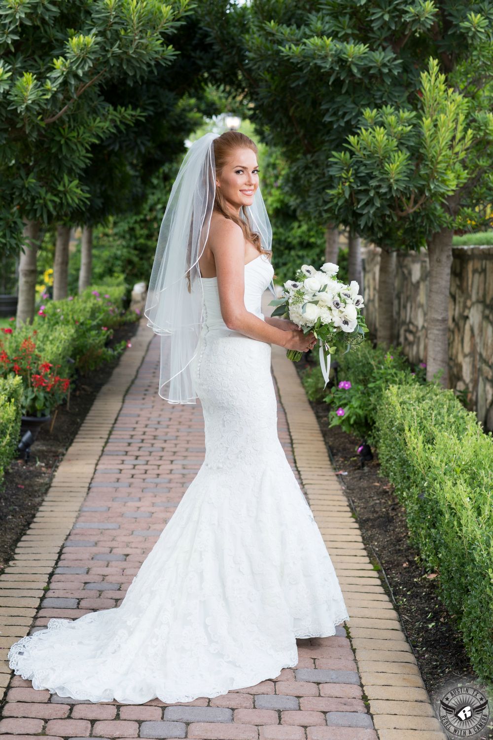 Color picture of gorgeous tall redhead bride in elegant white strapless gown stands with white bridal bouquet from Visual Lyrics Floral Art between tress at Nature's Point Austin wedding venue on the shores of Lake Travis on her wedding day taken by austin wedding photographer.