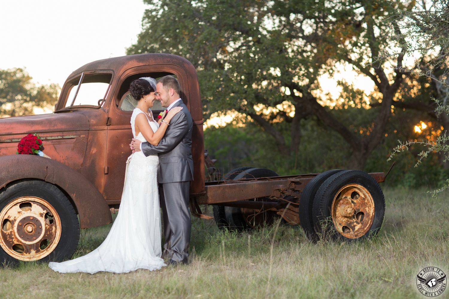 Austin wedding photgrapher takes picture of bride in white wedding gown with red rose bridal bouquet and groom in shiny grey suit embracing on their wedding day in front of rusted old truck at Star Hill Ranch Austin area wedding venue in the Hill Country.