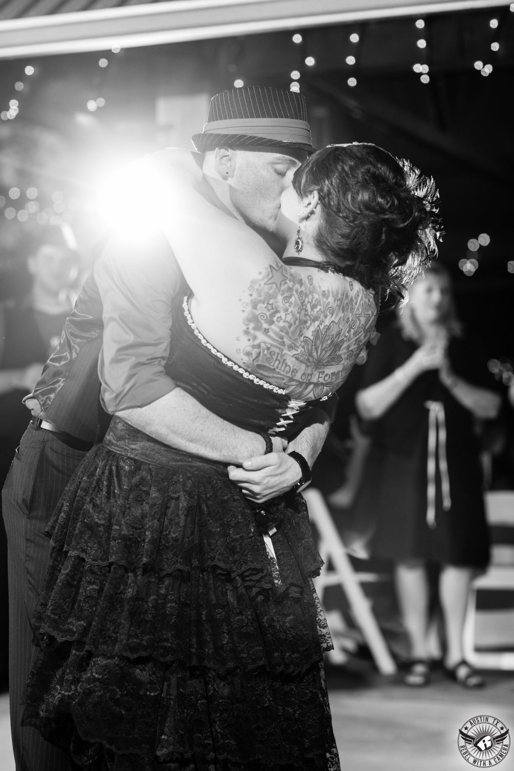 Dramatic black and white wedding picture of off beat bride in black lace dress and tattoos kisses groom in black suit and hat during their first dance at their wedding reception at Cedar Bend Events in the Austin, Texas, area taken by Austin wedding photographer.