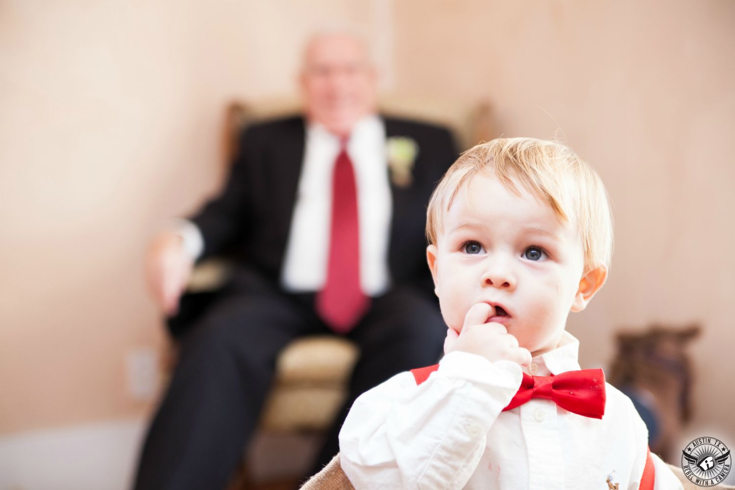Wedding picture of adorable ring bearer in red bowtie standing quizzically in front of grandpa with red tie and black suit in the bridal cottege at Star Hill Ranch Hill Country wedding venue taken by Austin wedding photogragrapher.