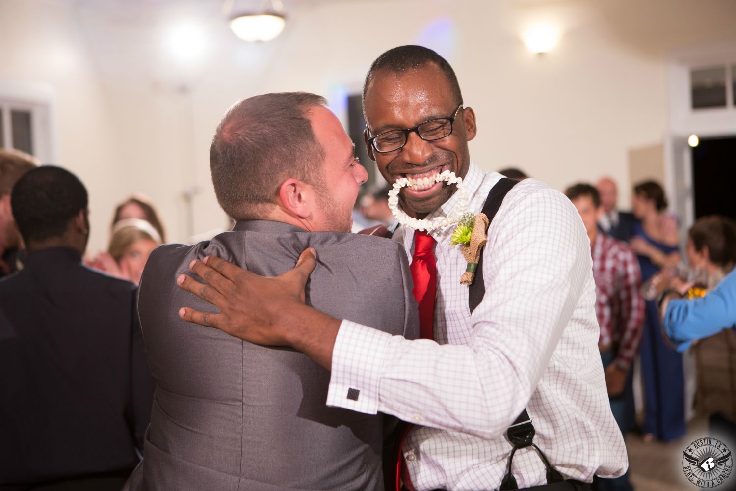 Picture taken after garter toss groom hugging best man who caught the garter and is holding it in his teeth at wedding reception at Star Hill Ranch wedding venue in the Austin area by Austin wedding photographer.