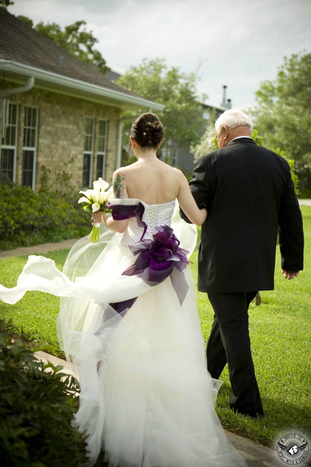 Austin wedding photographer took picture of back of bride in beautiful custom designed haute couture wedding dress with deep purple sash and her father in black tuxedo walking to the wedding ceremony at the Phoenix wedding venue in Columbus, Texas.