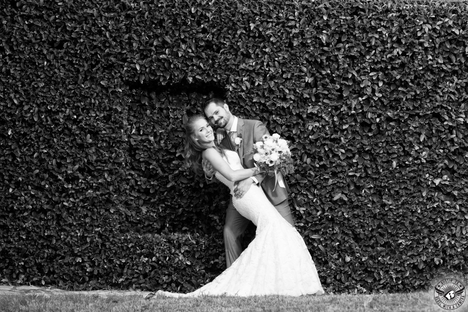 Black and white wedding picture of gorgeous red head bride in stunning, strapless, white wedding gown with exquisite white anemone bridal bouquet by Visual Lyrics Floral Art is being dipped by groom in grey suit in front of fig ivy covered wall on their wedding day at Nature's Point Austin area wedding venue.