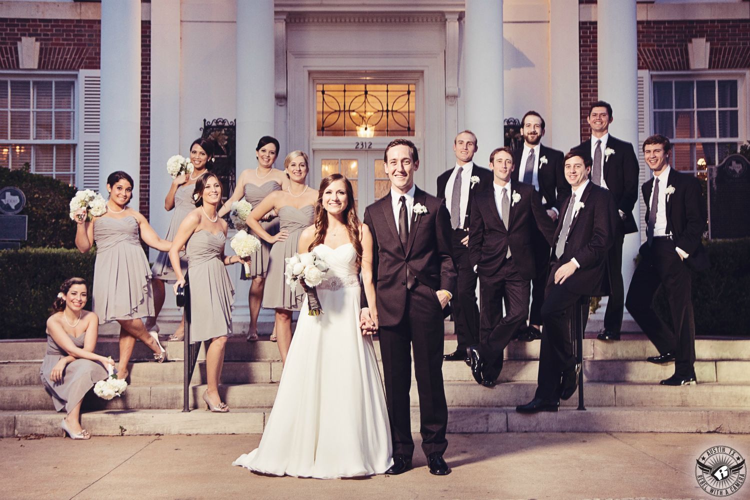 Wedding picture of entire bridal party with bridesmaids in gray dresses and groomsmen in black suits with black ties on the steps of the Texas Federation of Women's Clubs Mansion Headquarters Austin wedding venue in downtown Austin.