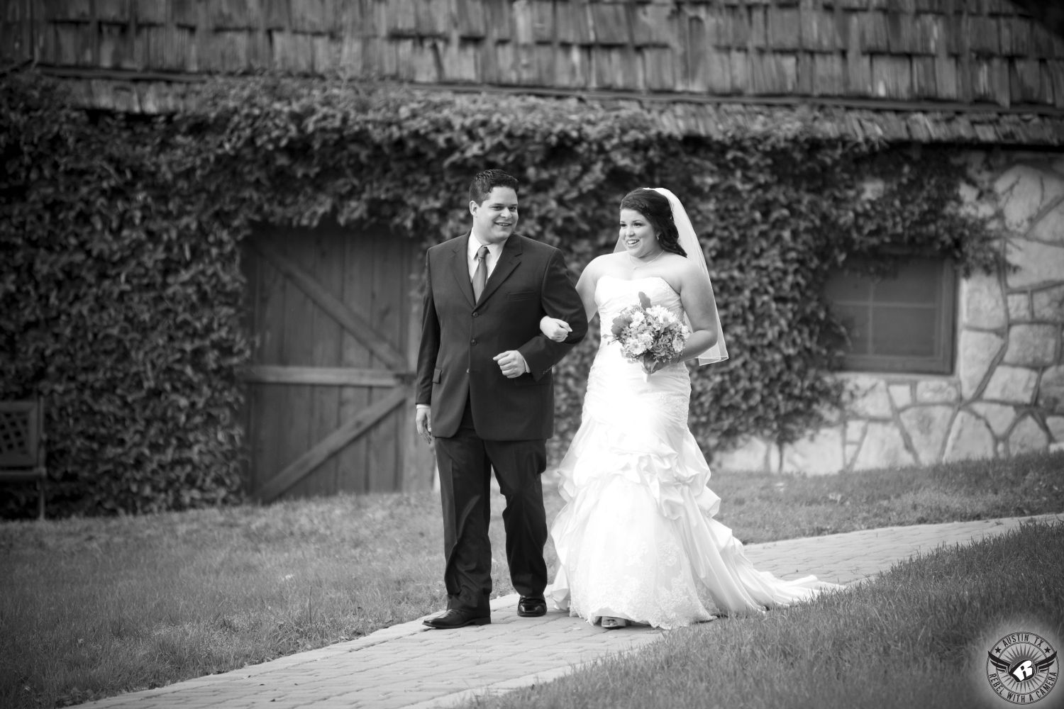 Black and white image of bride in strapless white wedding gown with lovely bouquet by Visual Lyrics Floral Artistry is escorted down the aisle by her brother in front of the gatehouse covered in fig ivy at Nature's Point to the arbor at the outdoor wedding ceremony site at Nature's Point Austin wedding venue on the shores of Lake Travis.