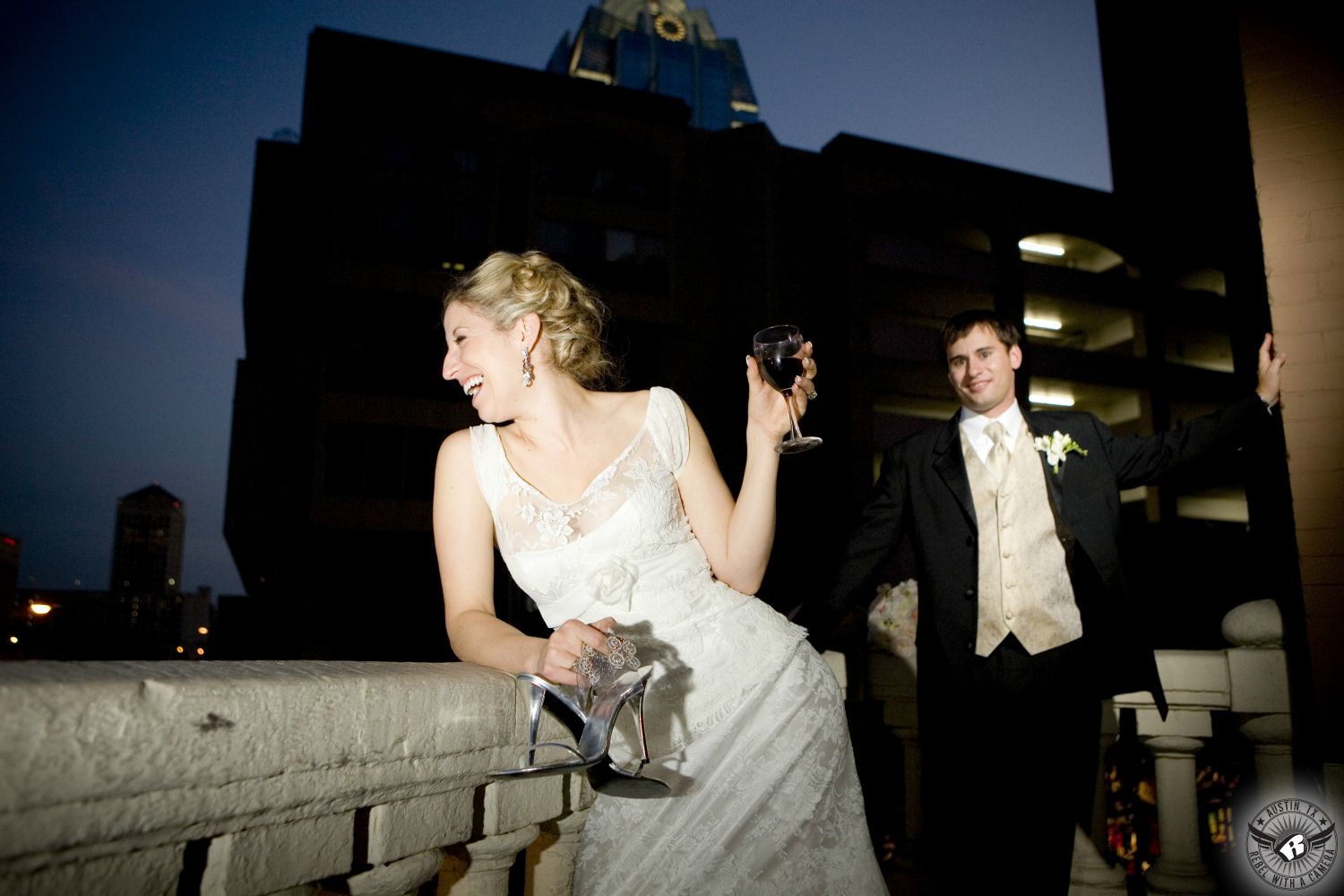 Happy blonde bride with makeup by Rhea McCarter Craft hold glass of red wine and her silver high heels with groom in black tuxedo and white vest and tie behind her on the balcony of the Driskill Hotel with the Frost Building in the background in downtown Austin on ther wedding day.