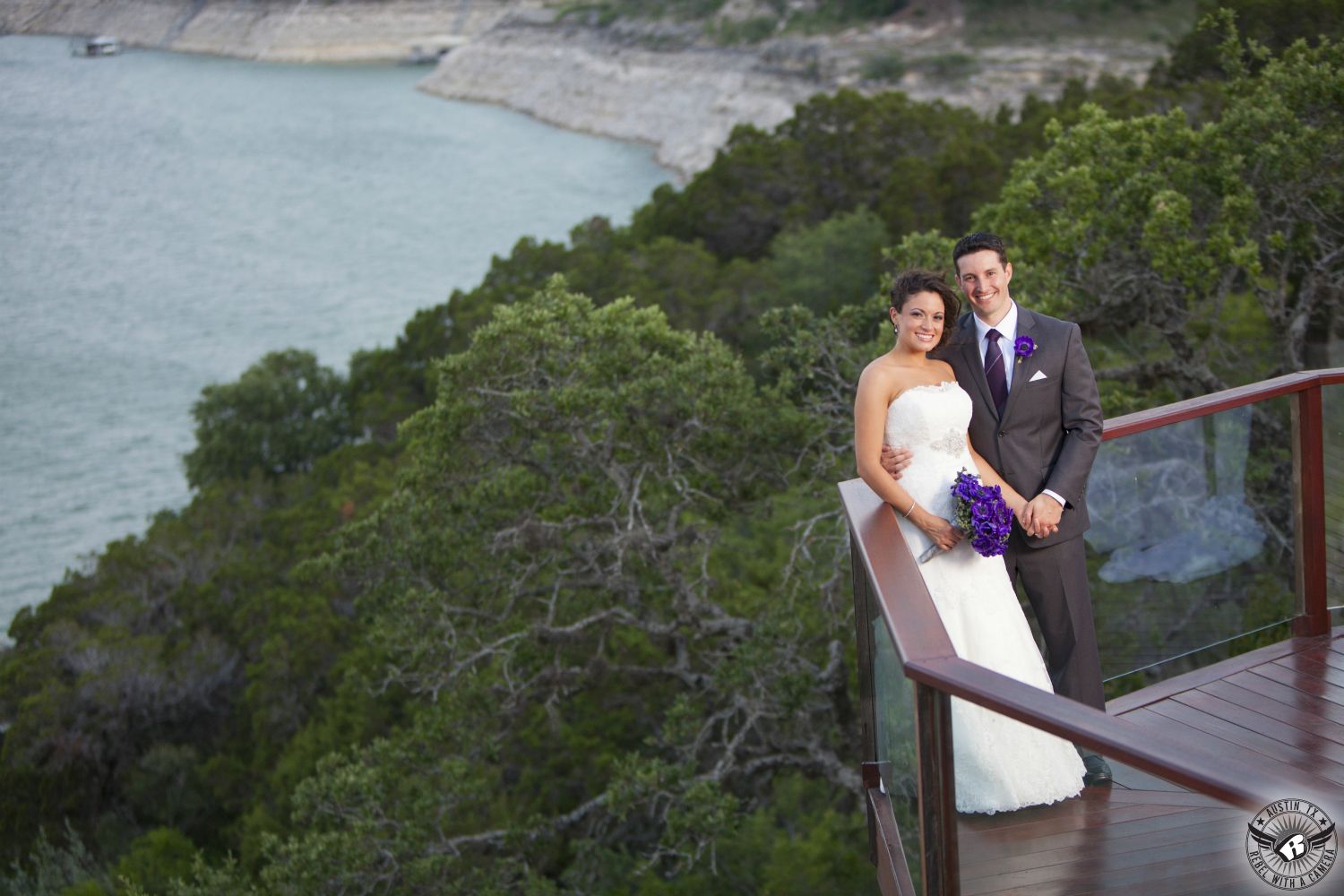 Bride in Priscilla of Boston strapless white lace wedding gown with sparkly sash and carrying bridal bouquet of purple anemone flowers by Visual Lyrics Floral Artistry and groom in grey suit and and eggplant colored tie with violet  anemone boutonniere on deck overlooking Lake Travis at Nature's Point Austin wedding venue.