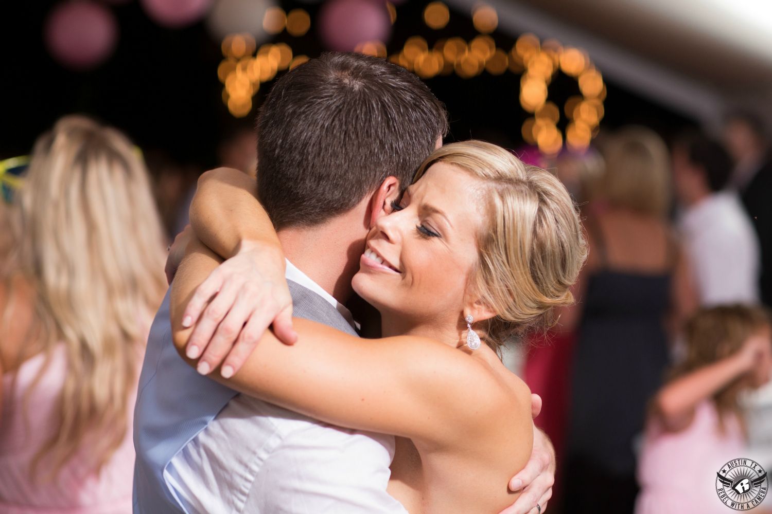 Smiling blonde bride in strapless wedding gown in dangly earrings hugs groom during their first dance at wedding reception under tent with twinkly lights in the background at Cedar Bend Events wedding venue south of Austin. 