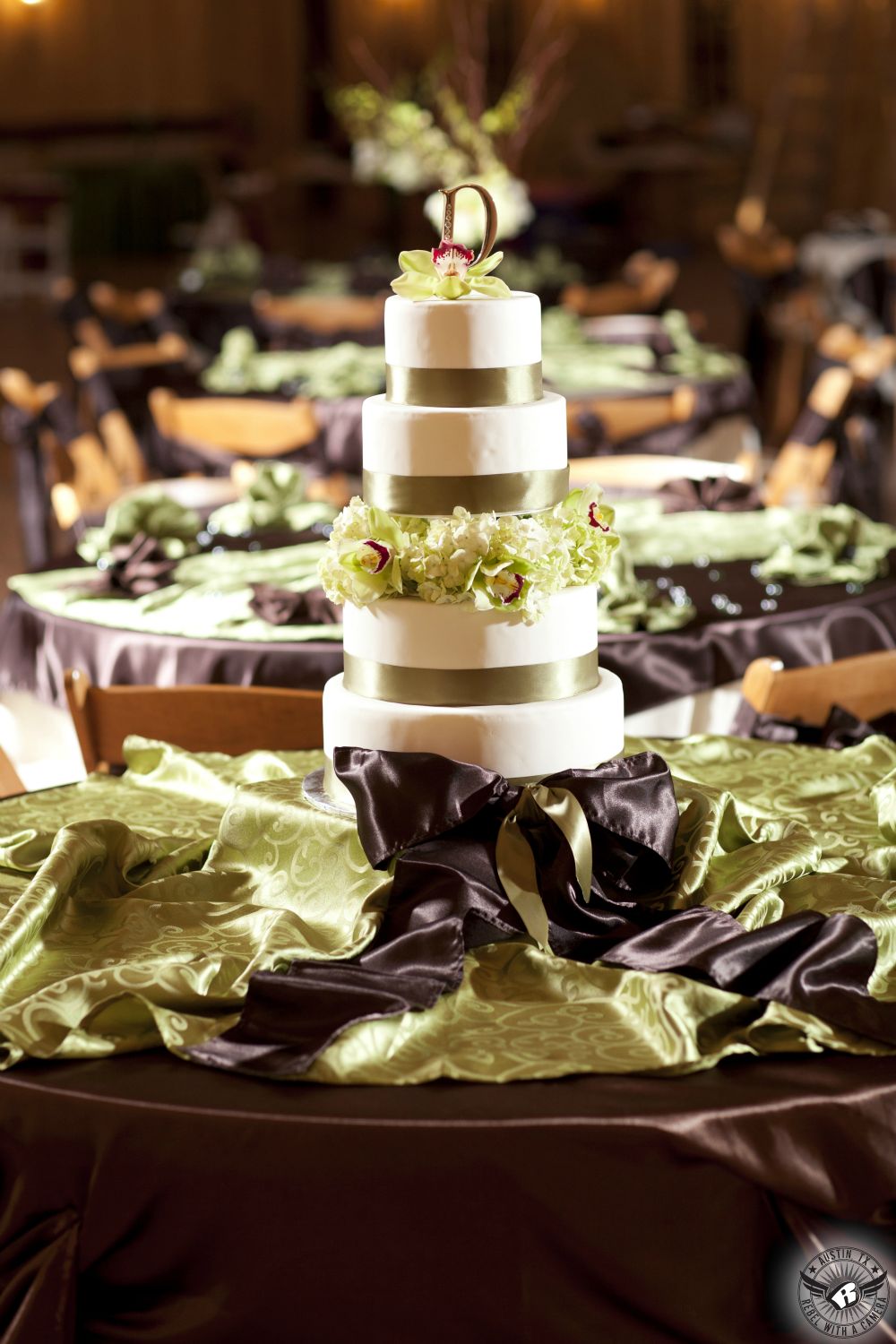 Picture of stunning four tier cake with Cymbidium orchids and hydrangea and light green ribbon on table with brown and sea green swirled linens at Gabriel Springs Austin area wedding venue by Austin wedding photographer.