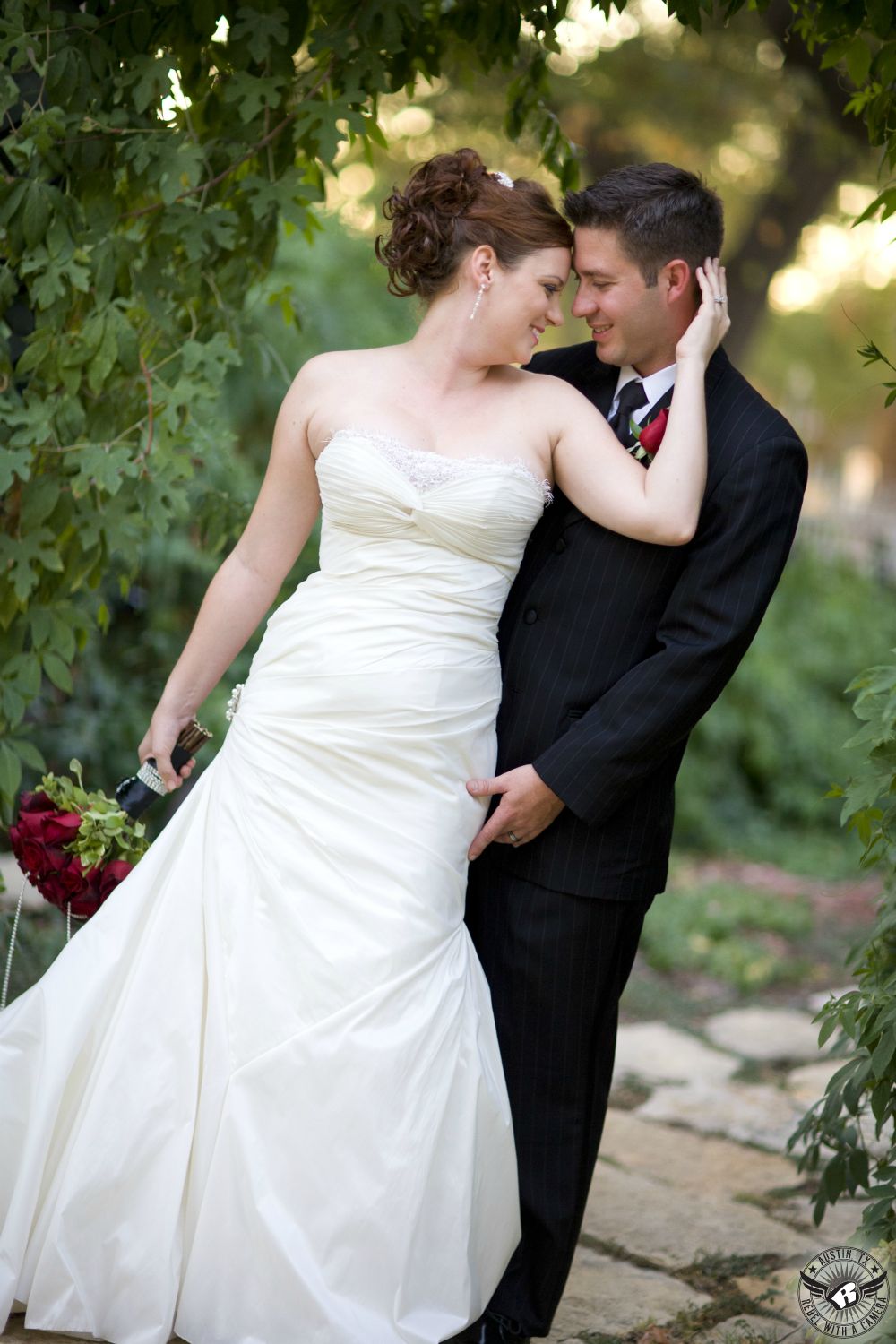 Bride with red rose bridal bouquet by Creative Innovations florist and groom in black tuxedo and red rose bud boutonniere embrace in front of vine covered arch at Inn at Salado Texas wedding venue.