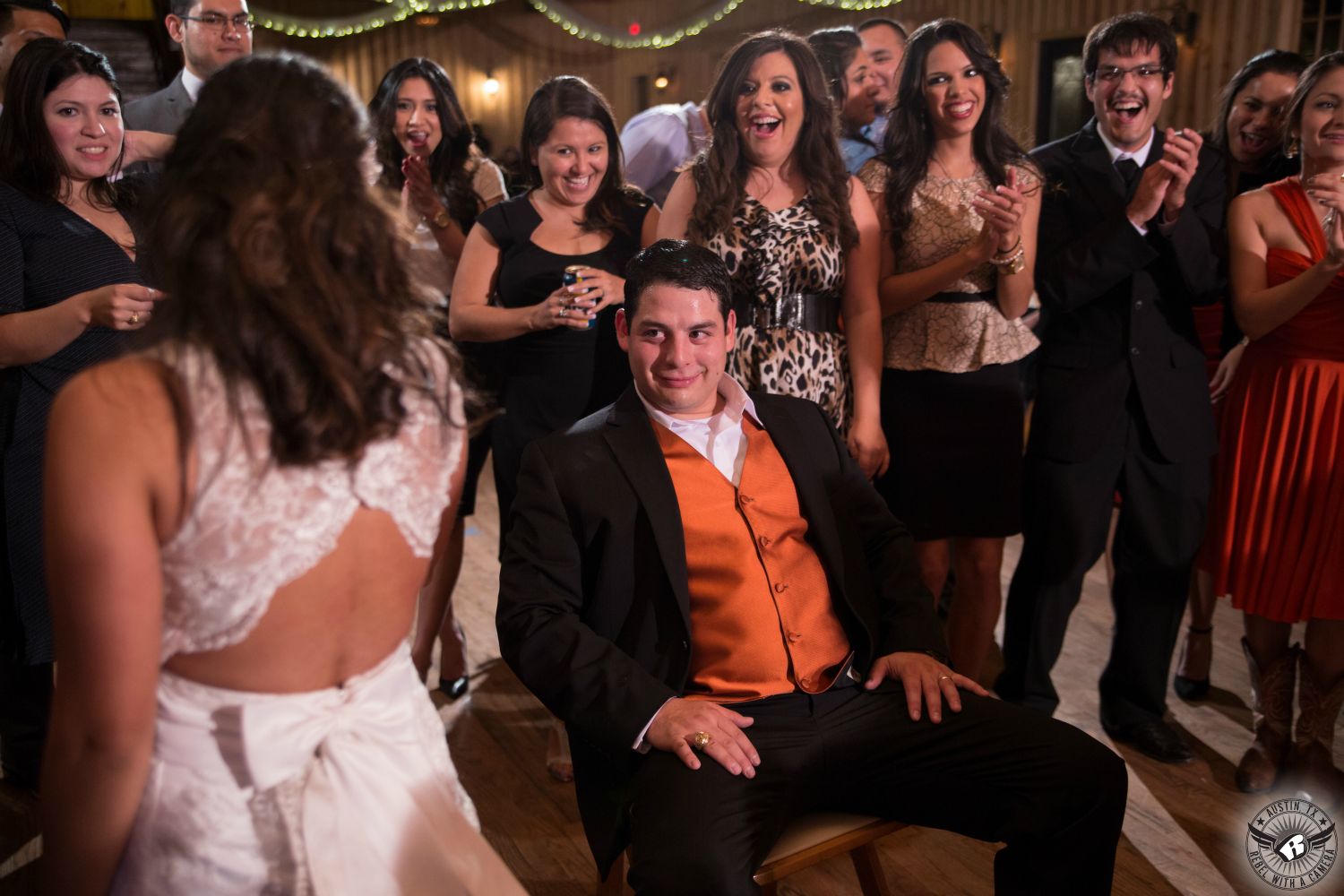 Groom in black tuxedo and orange vest sits in chair and looks at bride in backless wedding dress as she dances during wedding reception as guests look on and with twinkly lights in the background at Gabriel Springs austin area wedding venue.