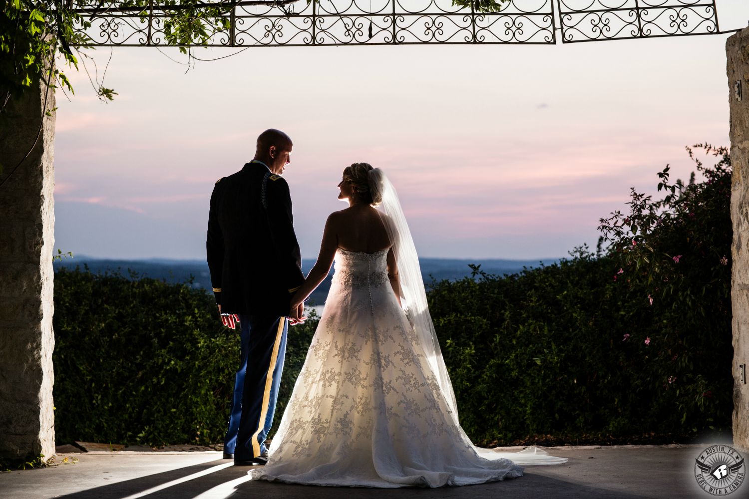 Dynamic backlit picture of silhouette of bride in white strapless lace dress and long veil and groom in army military dress blues in front of sunset at wedding ceremony arbor at Vintage Villas Austin wedding venue on Lake Travis taken by Austin wedding photographer.