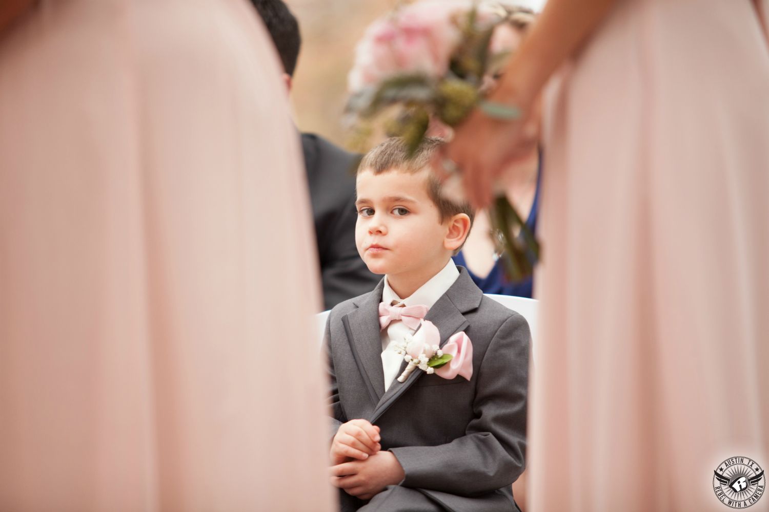 Wedding picture of adorable ringbearer in grey suit with light pink rose boutonniere and pink bowtie looks at the camera through bridesmaids holding beautiful pink bouquets by HEB Blooms during wedding ceremony at the arbor at Nature's Point Austin wedding venue.