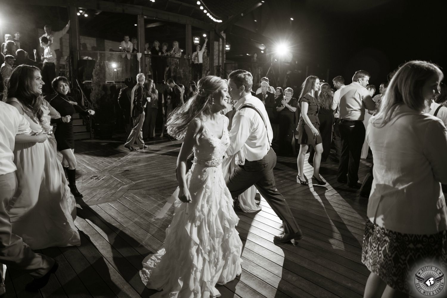 Bride in delicately ruffled bridal gown and groom in suspenders link arms and dance on the lower deck during their wedding reception at Nature's Point Austin wedding venue.