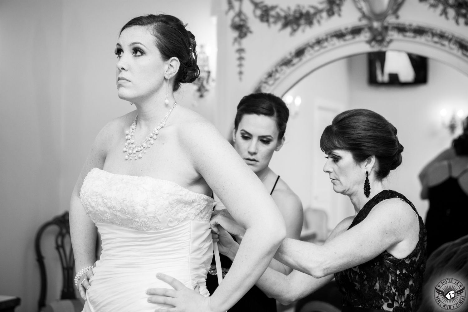 Dramatic black and white images of bridesmaids in black dresses lacing up corset of bride's wedding dress in front of large decorated mirror in bridal room at Villa St. Clair Austin wedding venue taken by Austin wedding photographer.
