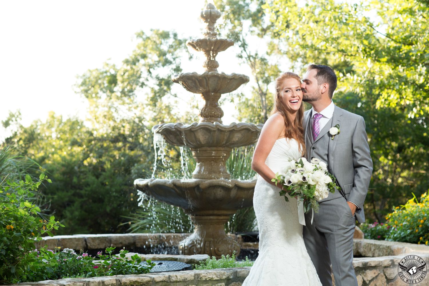 Groom in grey suit kisses gorgeous, tall, red headed bride in strapless bridal gown by beautiful white anemone bridal bouquet by Visual Lyrics Floral Artistry on the cheek in front on beautiful fountain at the Austin wedding venue Nature's Point.