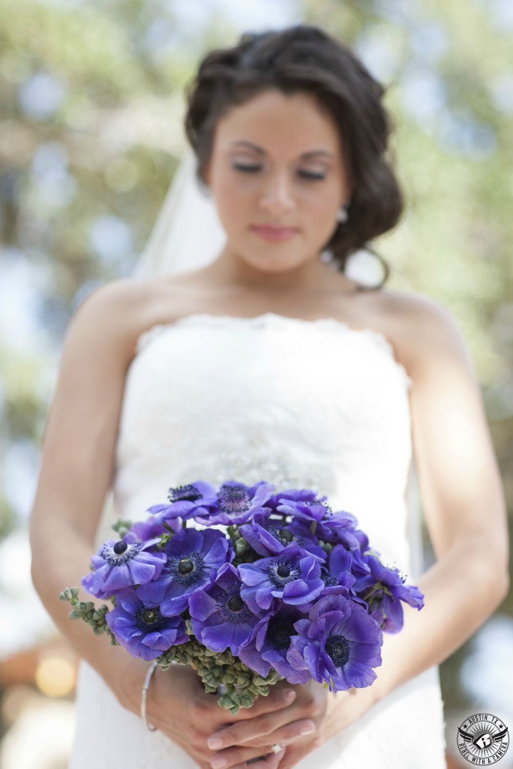 Gorgous brunette bride in white, strapless lace bridal gown by Priscilla of Boston holds and looks down at exquisite purple anemone bridal bouquet by the Austin florist Visual Lyrics Floral Artistry at Nature's Point Austin wedding venue.
