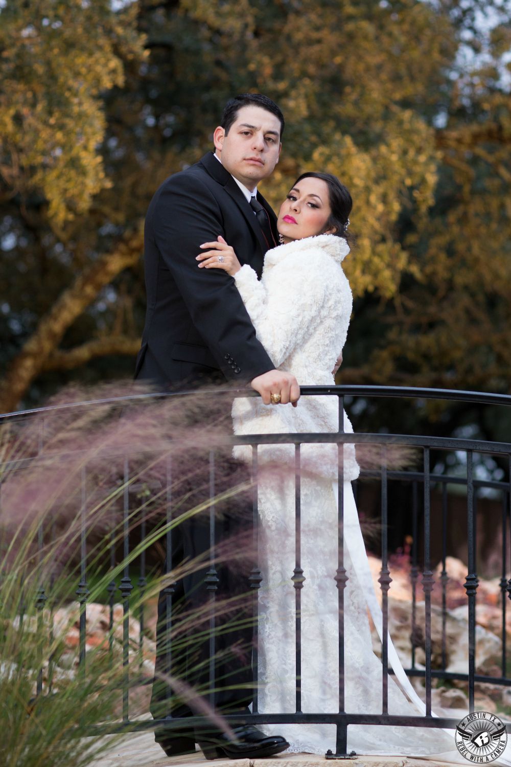 Glamorous and dramatic bride in white fur coat and groom in black tuxedo stand on bridge during winter wedding at Gabriel Springs wedding venue near Austin.